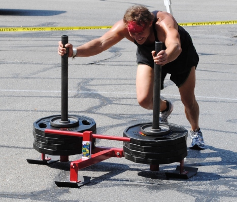 fit middle aged man in black workout clothes pushing sled full of weights in parking lot