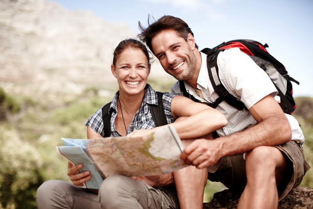 young couple in hiking gear sitting down smiling and reading a map with a mountain backdrop