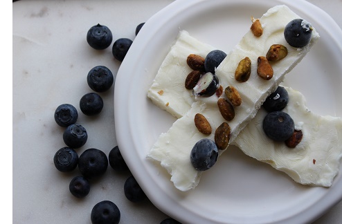 frozen piece of white yogurt encrusted with pistachios and blueberries overlapping another piece on a white plate with blueberries adjacent